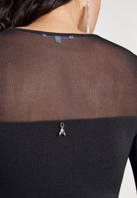 Top nero con un pannello trasparente sul retro, accentuato da un charm argentato. Texture liscia con scollatura rotonda e dettagli di cucitura sottili.