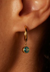 Goldene Creolen mit einem blauen Edelsteinanhänger, der eine runde Form und eine glatte Oberfläche aufweist. Der Edelstein hängt an einer kleinen Schlaufe.