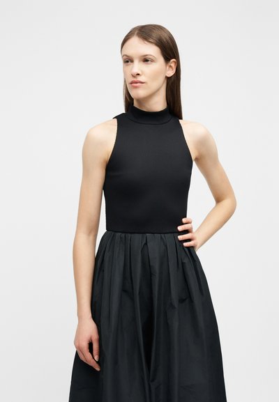 Jeune femme aux cheveux bruns raides portant un haut noir sans manches à col montant et une jupe noire taille haute, debout avec la main sur la hanche.