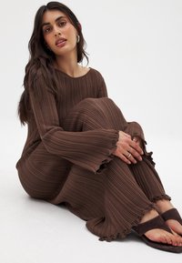 Robe en maille côtelée marron avec un décolleté large et des manches évasées, présentant des bords volants. Associée à des sandales à glisser marron.