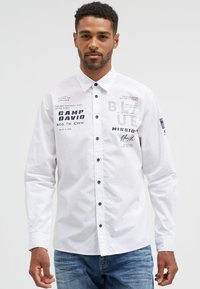 White button-up shirt with black accents and graphic prints, featuring various texts. Long sleeves with buttoned cuffs, made from cotton fabric.