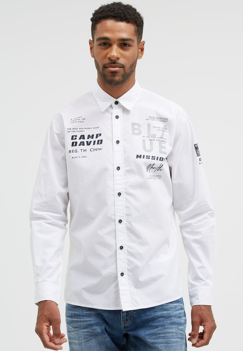 White button-up shirt with black accents and graphic prints, featuring various texts. Long sleeves with buttoned cuffs, made from cotton fabric.