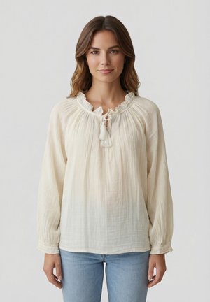 Giovane donna che indossa una blusa crema fluida con scollo arricciato e lacci con nappine, abbinata a jeans azzurri, in piedi davanti a uno sfondo semplice.