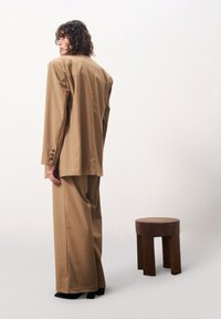 Brauner maßgeschneiderter Blazer und weite Hose, beide aus einem glatten Stoff, mit Knöpfen an den Manschetten und schwarzen Schuhen mit Blockabsatz. Dekorierter Hocker daneben.