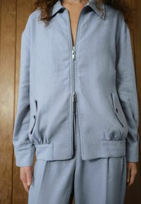 Light blue jacket made of a smooth fabric, featuring a zippered front, two side pockets, and elasticized waistband for fit.