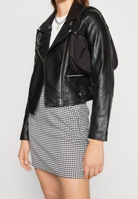 Schwarze Leder-Motorradjacke mit silbernen Reißverschlüssen, kombiniert mit einem schwarz-weiß karierten Minirock im Hahnentritt-Muster und einer kleinen schwarzen Tasche.