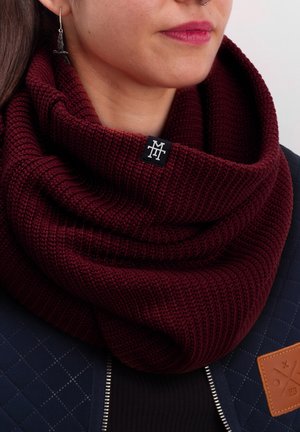 Burgundy knitted infinity scarf with a textured pattern, featuring a small black logo tag. Worn over a navy quilted jacket.