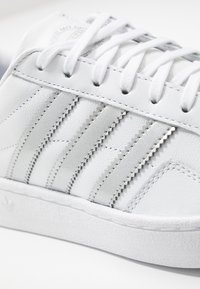 Vita lädersneakers med tre metalliska silverränder, platta vita snören och en texturerad gummisula. Närbild som framhäver sömnadsdetaljer.