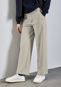 Pantalones de pierna ancha beige con una textura suave, que presentan bolsillos delanteros y un cierre de botón, combinados con zapatillas blancas.