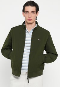 Geacă bomber verde cu fermoar frontal, două buzunare laterale și manșete canelate. Purtați peste un tricou polo cu dungi albastre deschise. Textură netedă a materialului.