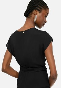Blusa nera con maniche corte e scollatura rotonda, caratterizzata da un dettaglio di chiusura sul retro e un design con vita legata. Texture liscia.
