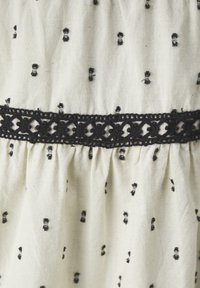 Cream fabric with black embroidered patterns resembling bows, featuring a black lace trim. Textured with gathers below the trim.
