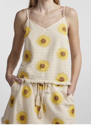 Kobieta ubrana w kremowy dziany top na ramiączkach i krótkie spodenki z żółtym wzorem słoneczników oraz ściągaczem w pasie, ręce w kieszeniach.