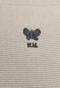 Papillon noir brodé et les lettres "FUU" sur un tissu tricoté beige.