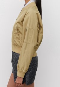 Giacca bomber color cammello con chiusura frontale a zip, polsini e orlo a coste, e una tasca con zip sulla manica, abbinata a pantaloni corti grigi trapuntati.