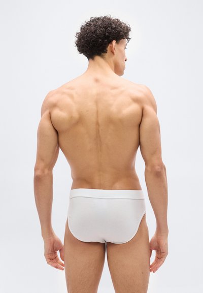 Μυώδης άνδρας με σγουρά μαλλιά, γυρισμένος προς τα πίσω, φοράει λευκά σλιπ σε απλό λευκό φόντο.