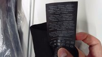 Ruka drží čiernu odevnú etiketu zobrazujúcu zloženie látky a symboly údržby v niekoľkých jazykoch na bielom pozadí.
