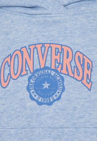 Svijetloplava dukserica s velikim narančastim natpisom "CONVERSE" i kružnim plavim logotipom s zvijezdom. Mekana tkanina, ležeran dizajn, rebrasti rubovi.