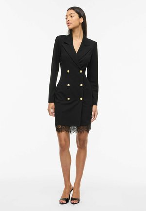 Žena v čiernych dvojradových šatách s golierom, zdobených zlatými gombíkmi a čipkovým lemom, v kombinácii s čiernymi sandálmi s otvorenou špičkou, stojaca na bielom pozadí.