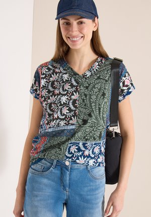 Junge Frau lächelt, trägt eine blaue Kappe, ein V-Ausschnitt-Hemd mit Patchwork-Blumenmuster, hellblaue Jeans und eine schwarze Umhängetasche.