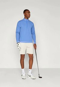 Pullover azzurro chiaro con zip, pantaloni corti beige, scarpe da ginnastica bianche, mentre tiene un bastone da golf, in piedi contro uno sfondo neutro.