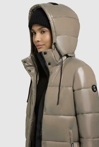 Giacca puffer di un taupe chiaro con finitura lucida, cappuccio staccabile, dettagli neri, tasche laterali e cuciture in vista. Beanie nero indossato.