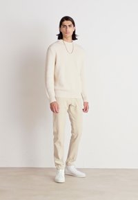 Pulover din crem deschis, cu model în riduri, și pantaloni bej, asortați cu adidași albi. Ținuta are o croială lejeră și texturi netede.