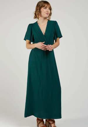 Femme portant une robe maxi vert foncé à col en V avec des manches courtes volantées, debout les mains jointes et regardant de côté.