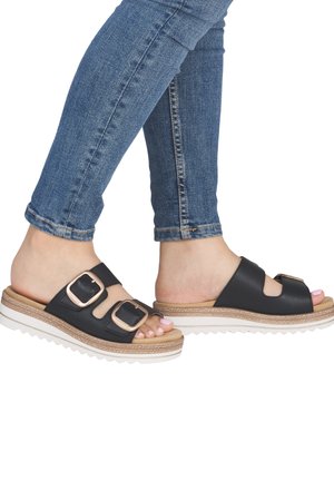 Pieds portant des sandales noires à enfiler avec deux boucles dorées, associées à un jean bleu ajusté sur fond blanc.