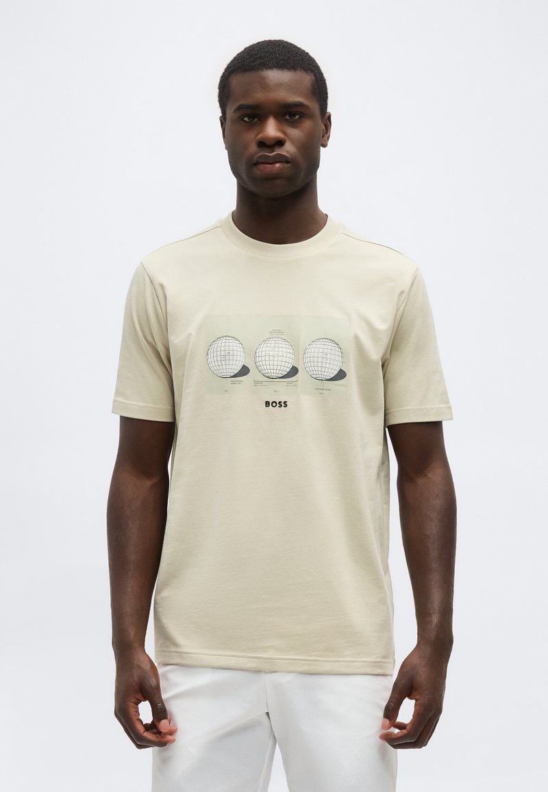 Άνδρας που φοράει μπεζ μπλουζάκι με τρεις απεικονίσεις υδρογείου και το κείμενο «BOSS», στέκεται μπροστά σε λευκό απλό φόντο.