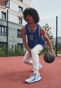 Basketbollspelare i en blå ärmlös tröja, vita byxor och ljusblå sneakers med marinblå detaljer, som dribblar en svart Spalding-basketboll på en plan.