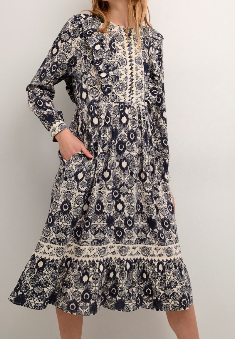 Femme portant une robe midi à manches longues bleu marine et blanche à motifs, avec des volants et des poches, debout devant un fond clair uni.