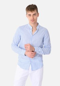 Camicia da uomo a maniche lunghe in blu chiaro e bianche a righe, con colletto alla mandarina e chiusura con bottoni, abbinata a pantaloni bianchi.