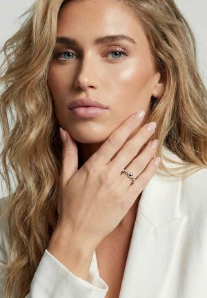 Lyshåret kvinde med bølget hår iført hvid blazer, der rører ved sin hage med venstre hånd og viser en sølvperlet ring og naturlig neglelak.