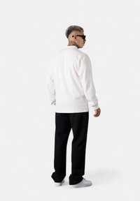 Weißer Sweatshirt mit langen Ärmeln, geripptem Kragen und lockerer Passform, kombiniert mit lockeren schwarzen Hosen und weißen Sneakers, von hinten betrachtet.