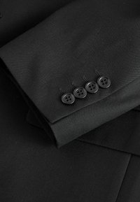 Manche de costume noire avec quatre boutons noirs disposés verticalement près du poignet, mettant en valeur la fine texture du tissu et les détails des coutures.