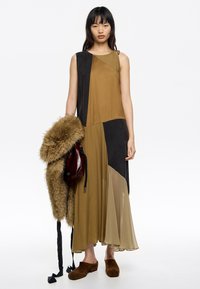 Maxi sukienka w brązowych odcieniach z kolorowymi blokami; posiada przezroczysty dół i jedno ramiączko. Modelka trzyma torbę z futrzanym akcentem.