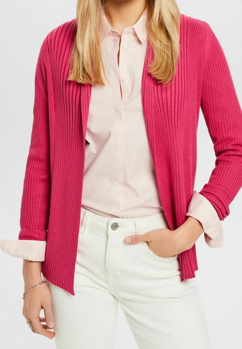Cardigan long en maille côtelée de couleur rose vif, avec une ouverture sur le devant et des manches longues. Assorti à une chemise rose clair à boutons et un jean blanc.