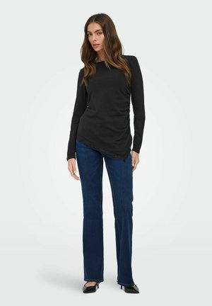 Kobieta ubrana w czarny top z długim rękawem, asymetrycznym dołem i koronkową lamówką, ciemnoniebieskie proste jeansy i czarne szpiczaste szpilki.