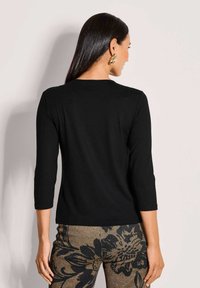 Schwarzes 3/4-Ärmel-Oberteil mit rundem Halsauschnitt und glatter Textur. Das Model trägt gemusterte Hosen mit floralen Designs in Brauntönen und Schwarz.