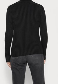 Schwarzer gerippter Pullover mit langen Ärmeln und hohem Kragen, der eine enganliegende Passform und strukturierten Stoff aufweist. Getragen mit dunkelgrauen Hosen.