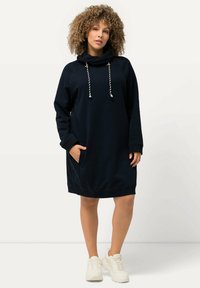 Navy Kapuzenkleid mit langen Ärmeln, Seitentasche und kontrastierenden Kordeln. Weicher Stoff mit lockerer Passform. Getragen mit weißen Sneakers.