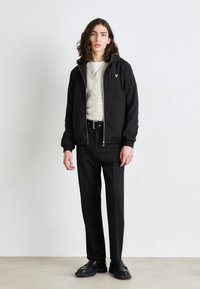 Jachetă neagră cu glugă, pulover tricotat gri, pantaloni negri. Prezintă un închidere cu fermoar și un accent cu logo. Textură netedă, design casual.