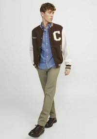 Veste de varsity marron et blanche avec un patch "C", chemise en denim bleue, pantalon vert olive et chaussures en cuir marron. Tenue décontractée avec des textures superposées.