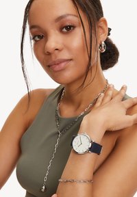 Marineblaues Armbanduhr mit einem silbernen Gehäuse und einem cremigen Zifferblatt, am Handgelenk getragen. Verfügt über ein glattes Zifferblatt und ein minimalistisches Design. Silberne Kettenkette sichtbar.