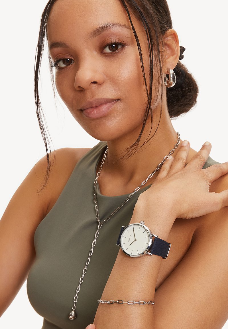 Marineblaues Armbanduhr mit einem silbernen Gehäuse und einem cremigen Zifferblatt, am Handgelenk getragen. Verfügt über ein glattes Zifferblatt und ein minimalistisches Design. Silberne Kettenkette sichtbar.