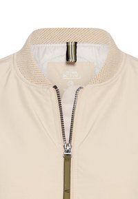 Veste beige avec col en maille côtelée, fermeture éclair argentée partiellement ouverte et détail de boucle rayée à l'intérieur du col.
