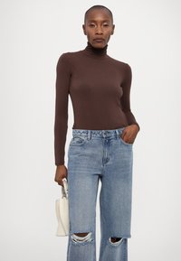 Maglione a manica lunga marrone con collo a roulè abbinato a jeans chiari a vita alta e strappati. La modella tiene in mano una piccola borsa bianca.