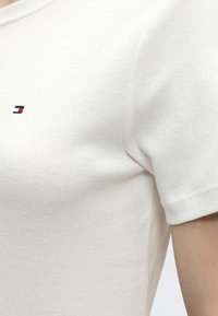 Κοντινή λήψη άσπρου βαμβακερού μπλουζάκι με κοντά μανίκια, με μικρό κόκκινο, άσπρο και ναυτικό ορθογώνιο λογότυπο στο στήθος κοντά στον ώμο.