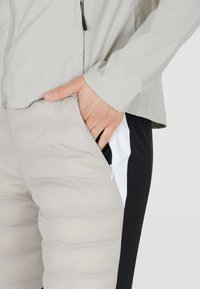 Ljust grå zip-jacka med en slät textur, kombinerad med beigefärgade och svarta byxor som har en dragkedjeförsedd sidficka och vita detaljer.